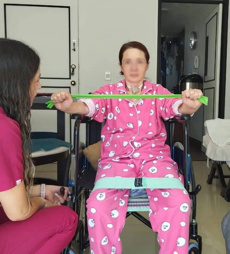 Valentina guiando ejercicios con bandas elásticas a una paciente en silla de ruedas, en su casa